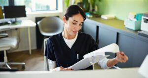 A woman in medical scrubs sits at a desk, looking down at papers in her hand. Office furniture, a window, and green walls are visible in the background, indicating a clinical or healthcare setting.