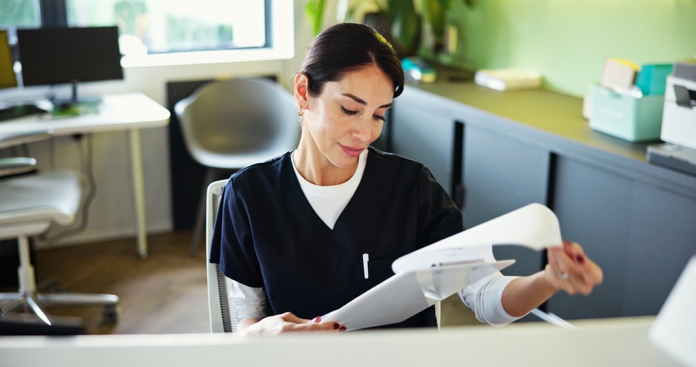 A woman in medical scrubs sits at a desk, looking down at papers in her hand. Office furniture, a window, and green walls are visible in the background, indicating a clinical or healthcare setting.